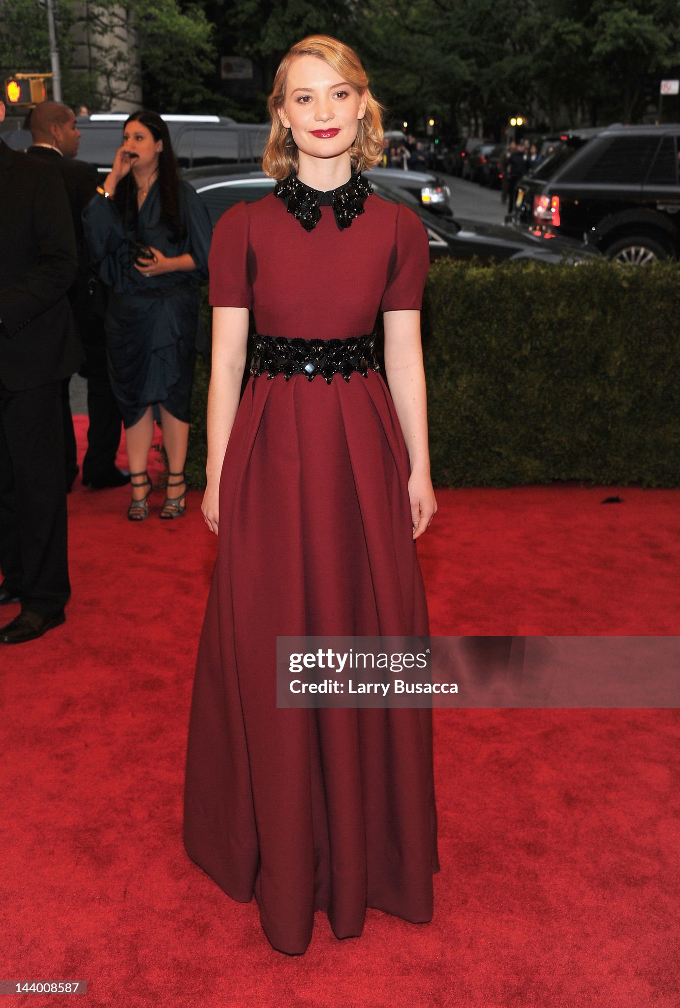 red carpet photo of slender white woman in floor-length burgundy gown, made of some stiff material, flat bodice with rounded shoulder and short sleeves, pleated skirt without much volume, sparkly black pointed collar and sash, with black clutch, blond hair in neat shoulder-length curls reminiscent of a 1940's schoolgirl, clean eyes, red lip